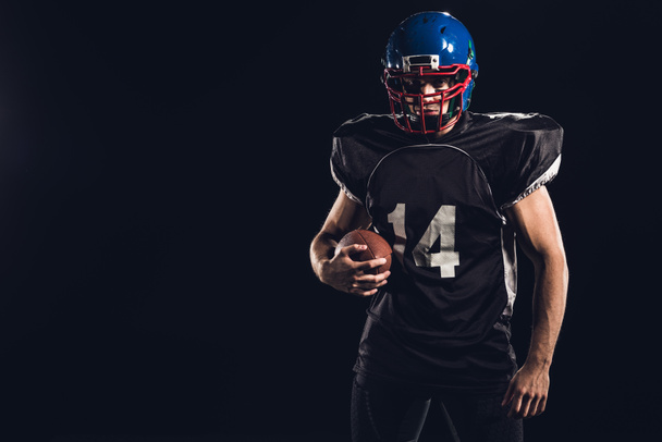 mladý americký fotbalista s míčem, při pohledu na fotoaparát izolované na černém pozadí - Fotografie, Obrázek