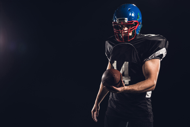 guapo jugador de fútbol americano con bola mirando a la cámara aislado en negro
 - Foto, imagen
