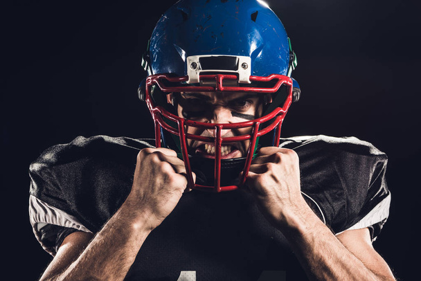 Close-up portret van boos Amerikaans football-speler in de helm camera geïsoleerd op zwarte kijken - Foto, afbeelding