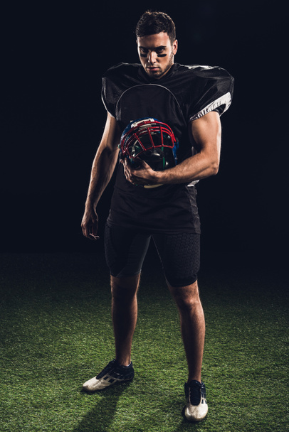 sérieux joueur de football américain en uniforme noir tenant casque et regardant la caméra sur noir
 - Photo, image