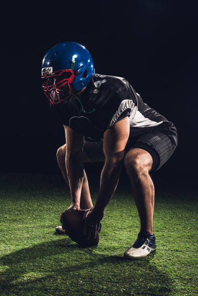 american football player standing squats on grass with ball between feet on black - 写真・画像