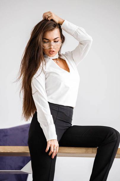 Studio portrait of young brunette woman in white shirt and black pants. Beauty business woman sitting on table at office interior - 写真・画像