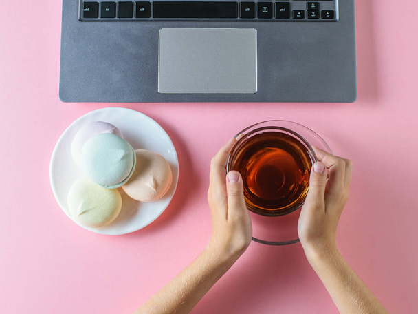 Девушка ест голубой зефир с чаем перед компьютером на розовом столе. Вид с вершины. Концепция здорового питания в современном мире. Пастельный цвет
. - Фото, изображение