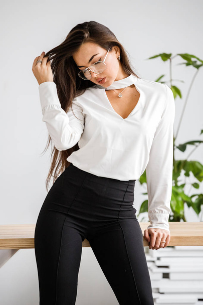 Studio portrait of young brunette woman in white shirt and black pants. Beauty business woman sitting on table at office interior - Foto, Bild