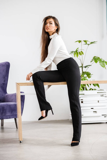 Studio portrait of young brunette woman in white shirt and black pants. Beauty business woman sitting on table at office interior - Valokuva, kuva