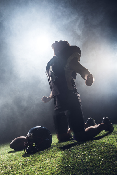 joven jugador de fútbol americano deprimido mirando hacia arriba y gritando mientras está de pie de rodillas sobre hierba verde contra el humo blanco
 - Foto, imagen