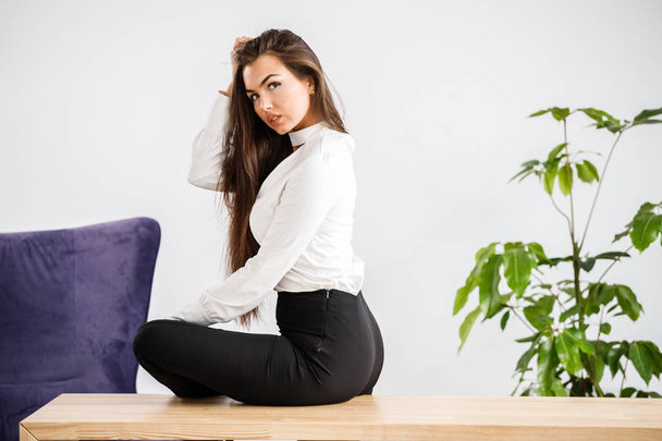 Studio portrait of young brunette woman in white shirt and black pants. Beauty business woman sitting on table at office interior - Photo, Image