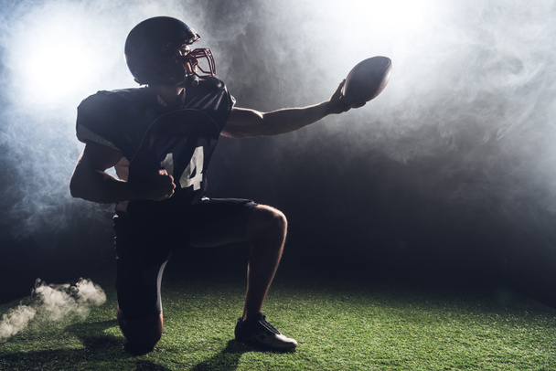 American football speler permanent op knie op groene gras en houden bal tegen witte rook - Foto, afbeelding