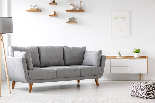 Simple, gray sofa standing next to a white cupboard in living room interior with decorations on wooden shelves. Real photo - Valokuva, kuva
