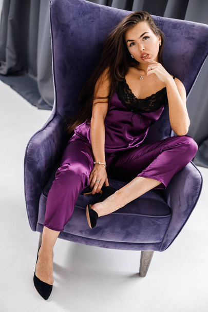 Studio portrait of young brunette woman in purple pajamas. Beauty woman sitting in violet armchair at home interior - Fotó, kép