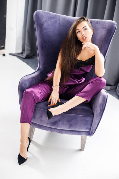 Studio portrait of young brunette woman in purple pajamas. Beauty woman sitting in violet armchair at home interior - Fotoğraf, Görsel