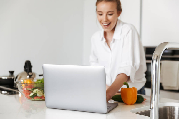 Cheerful young girl cooking salad while standing on a kitchen with laptop computer - Photo, Image
