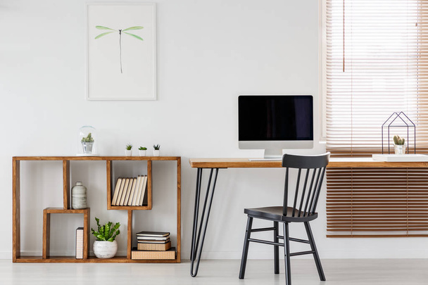 Real photo of a computer with empty screen standing on a wooden desk in bright freelancer's office interior with black chair and bookcase - Foto, Imagen