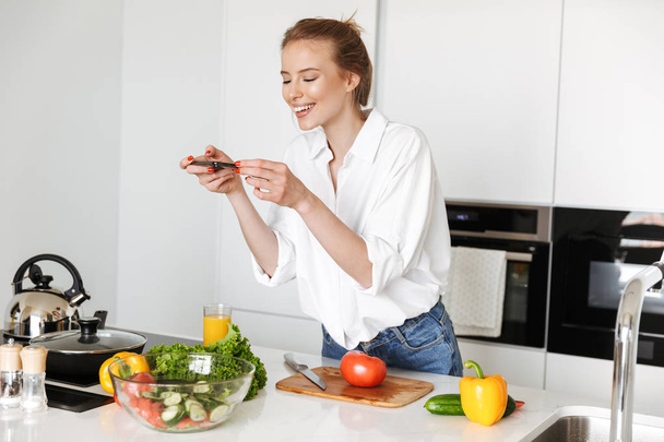 Happy young woman taking picture with mobile phone while cooking salad on a kitchen - Fotó, kép