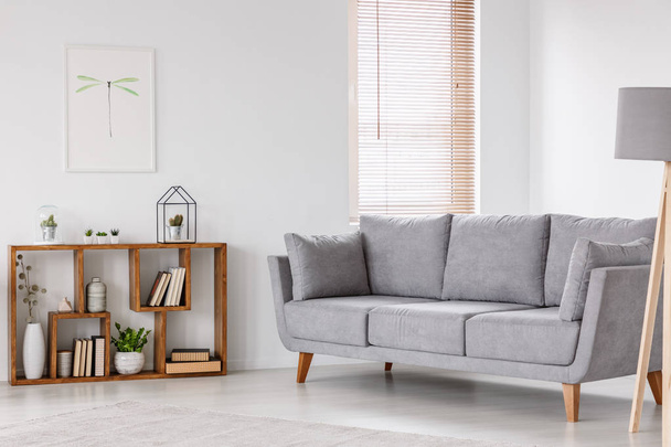 Real photo of a scandi living room interior with gray settee standing near the window, next to a wooden bookcase with plants and books - Zdjęcie, obraz