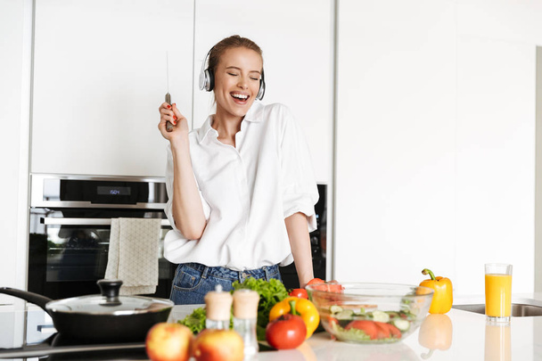 Cheerful young woman listening to music with headphones while cooking salad with knife in hand on a kitchen - Foto, immagini