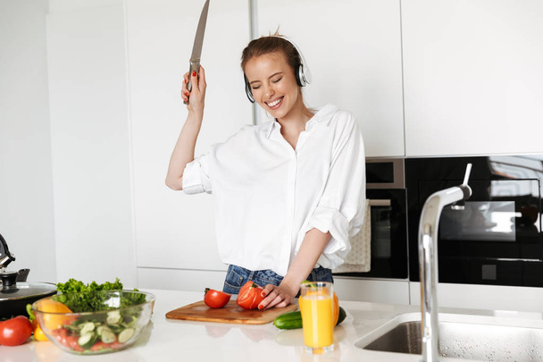 Iloinen nuori nainen kuuntelee musiikkia kuulokkeilla ruoanlaitossa salaatti veitsi kädessä keittiössä
 - Valokuva, kuva