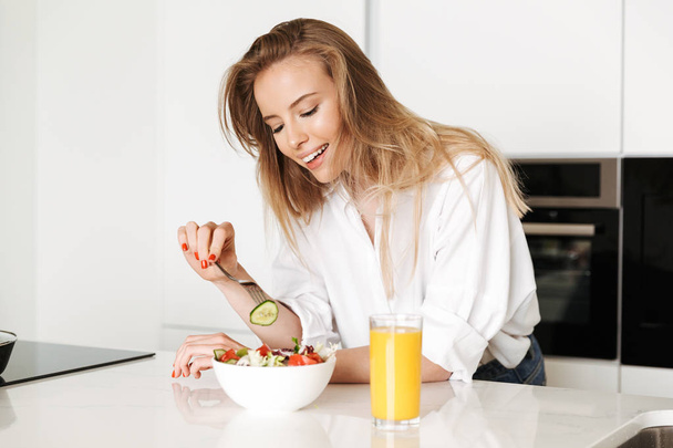 Smiling young woman eating salad from a bowl and drinking orange juice while standing on a kitchen - Фото, зображення