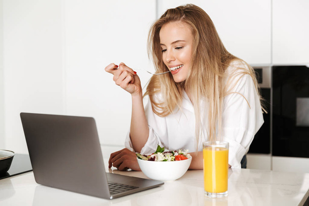 Cheerful young woman eating salad from a bowl and drinking orange juice while standing on a kitchen and watching movie on laptop - Photo, image