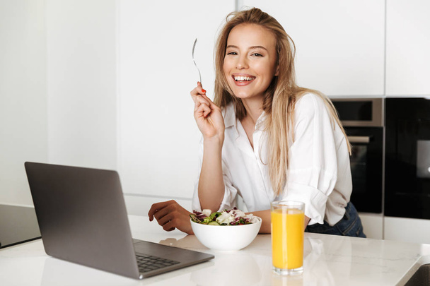 Satisfied young woman eating salad from a bowl and drinking orange juice while standing on a kitchen and watching movie on laptop - Photo, image