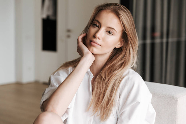 Beautiful young woman dressed in white shirt sitting on a couch at home and looking at camera - Фото, зображення