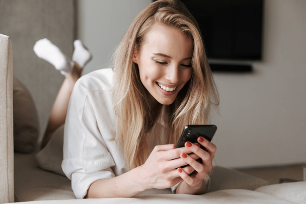 Cheerful young woman dressed in white shirt using mobile phone while laying on a couch at home - Foto, Bild