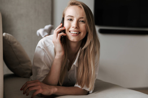 Photo of cheerful cute young woman indoors at home talking by mobile phone. - Φωτογραφία, εικόνα