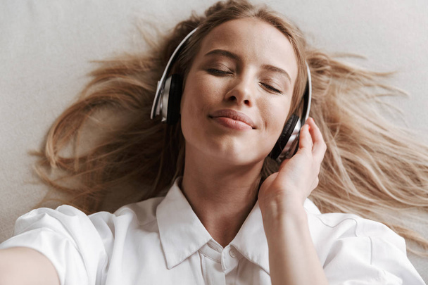 Top view of satisfied young woman in headphones taking a selfie while listening to music on a couch with eyes closed - Fotó, kép
