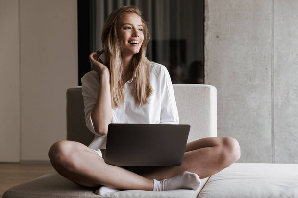 Happy young woman dressed in white shirt holding laptop computer while relaxing on a couch at home - Foto, Imagem