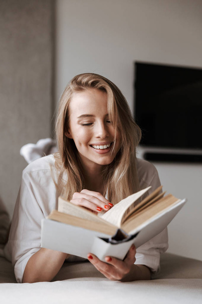 Happy young woman reading blank cover open book while relaxing on a couch at home - Фото, изображение