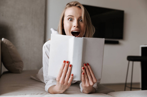Photo of cute shocked beautiful young woman indoors at home reading book. - Photo, image