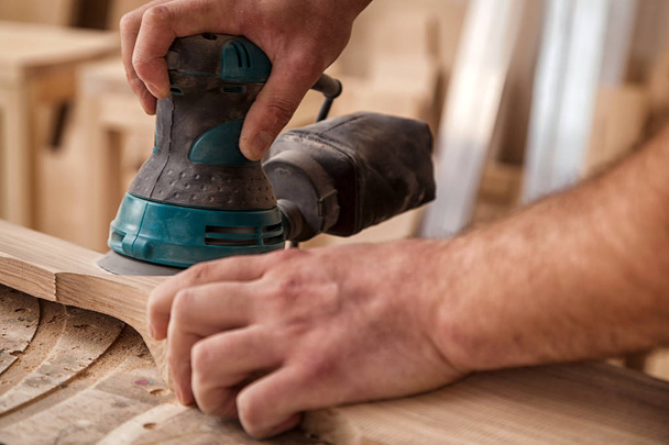 Крупным планом молодого плотника-строителя приравнивается полировка деревянной доски со случайным орбитным песком в мастерской
 - Фото, изображение