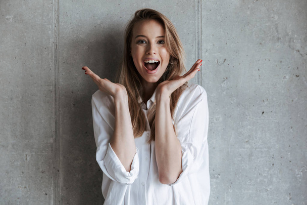Cheerful young woman dressed on white shirt standing over gray wall background with mouth open - Foto, Bild