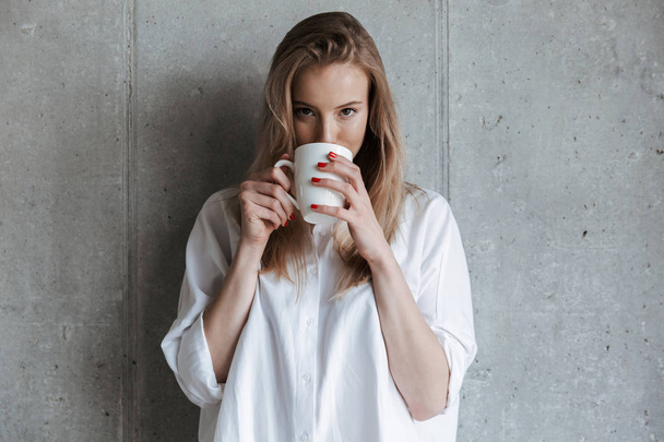 Picture of cute young woman indoors over grey wall drinking coffee holding cup. Looking camera. - Foto, Bild