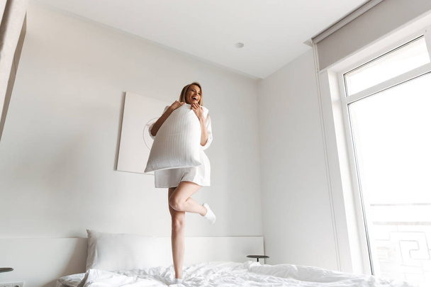 Фото молодой счастливой женщины, прыгающей с подушкой на кровати в помещении дома
. - Фото, изображение