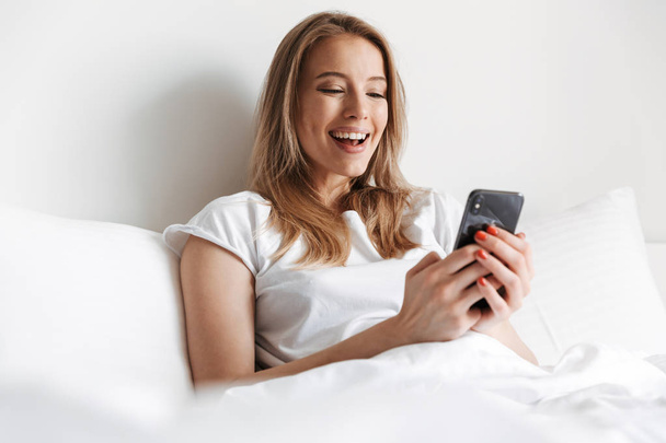 Photo of cute beautiful young woman indoors at home using mobile phone chatting. Looking aside lies in bed. - Foto, Imagen
