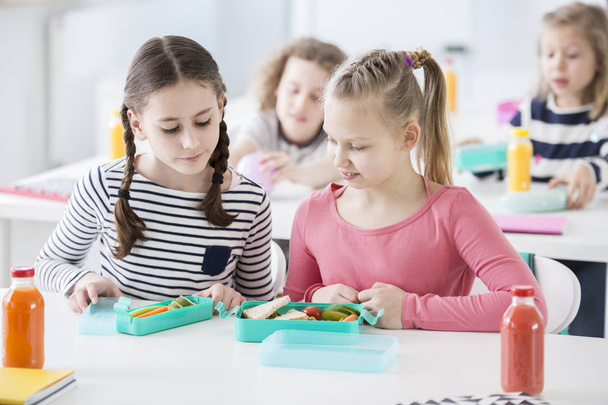 Two young girls during snack time in a school looking into each other's lunch boxes with healthy vegetables and bread. Bottles of fruit juices on the desk. Other kids in blurred background - Foto, imagen