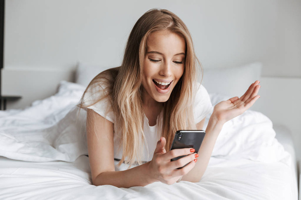 Photo of cute excited beautiful young woman indoors at home using mobile phone chatting. Looking aside lies in bed. - 写真・画像