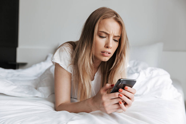 Photo of young sad woman lies on bed indoors at home using mobile phone. Looking aside. - Foto, imagen
