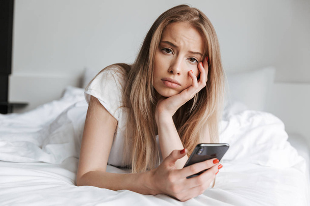 Photo of young sad woman lies on bed indoors at home using mobile phone. Looking camera. - Photo, image