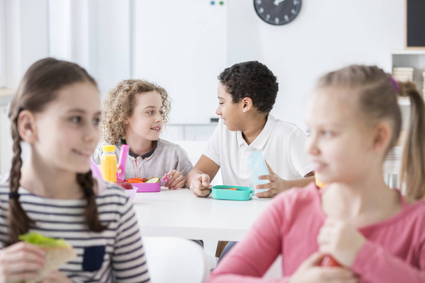 Two boys in the background talking during their lunch break in a school's classroom. Girls in the front, blurred. - Foto, imagen