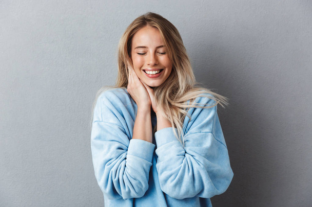 Portrait of a smiling young girl in blue sweatshirt with eyes closed isolated over gray background - Foto, Imagem