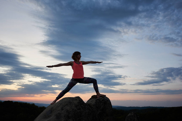 Young slim attractive woman doing gymnastic yoga exercises high on big rock on right blue pink evening sky at sunset background. Sport, fitness, health and active lifestyle concept. Virabhadrasana - Photo, Image