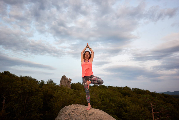 Slim fit jongedame doen yoga oefeningen staande blote voeten op een been op grote berg rots op groene bos tops achtergrond op felle zomerdag. Fitness en actieve levensstijl concept. Vrikshasana - Foto, afbeelding