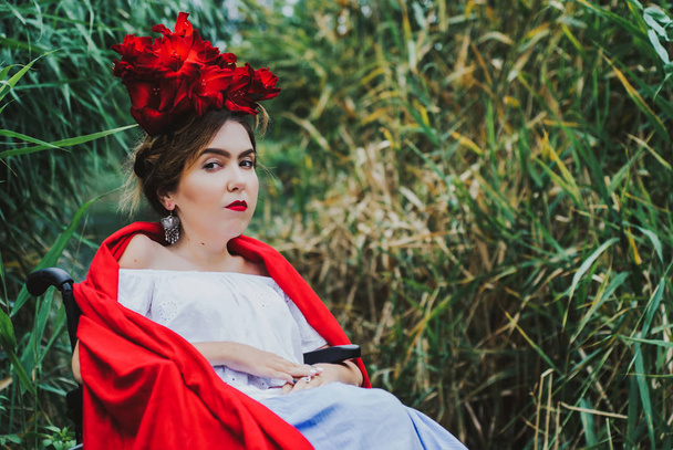 Summer conceptual portrait of beautiful young caucasian woman with red lips, red flowers in hair, sitting in wheel chair. frame with leaves wall on background. - Фото, зображення