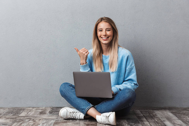 Smiling young blonde girl using laptop computer while sitting with legs crossed and pointing away isolated over gray background - Φωτογραφία, εικόνα