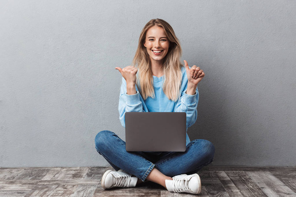 Smiling young blonde girl using laptop computer while sitting with legs crossed and pointing both ways isolated over gray background - Φωτογραφία, εικόνα