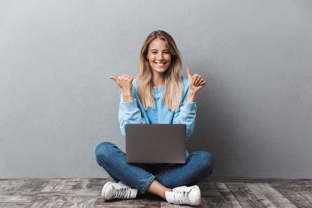 Happy young blonde girl using laptop computer while sitting with legs crossed and pointing both ways isolated over gray background - Φωτογραφία, εικόνα