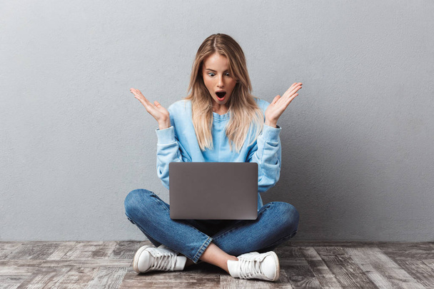 Amazed young blonde girl using laptop computer while sitting with legs crossed isolated over gray background - Φωτογραφία, εικόνα