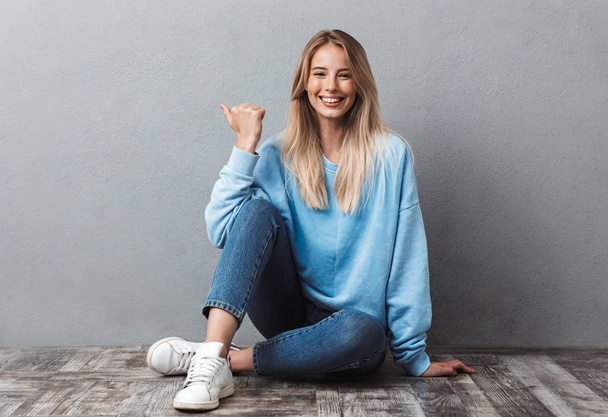 Happy young blonde girl pointing finger at copy space while sitting on a floor and looking at camera isolated over gray background - Фото, изображение